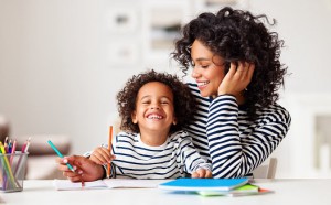 Cheerful ethnic woman laughing while helping happy boy to do homework assignment in cozy room at home